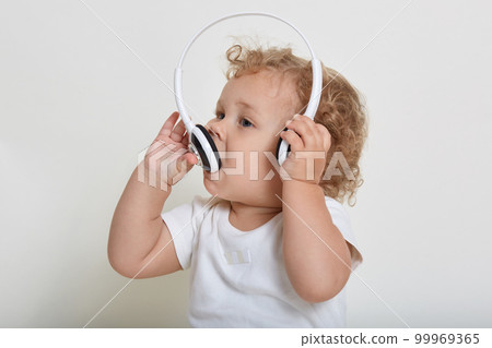 Charming baby boy with blond hair playing with headphones, biting it, wearing white t shirt, looking away, curious kid having fun, listening music. Charming baby boy with blond hair playing with headphones, biting it, wearing white t shirt, looking away, curious kid having fun, listening music. 99969365