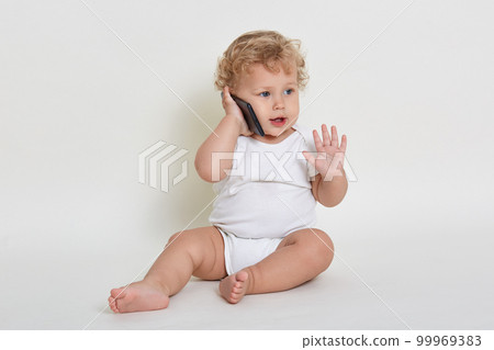Cute and joyful baby boy playing talking with somebody via smartphone while sitting indoors on floor, looking away and raising palm up, wearing white body suit, play with mobile phone. Cute and joyful baby boy playing talking with somebody via smartphone while sitting indoors on floor, looking away and raising palm up, wearing white body suit, play with mobile phone. 99969383