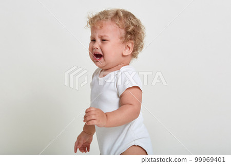 Cute baby boy standing against white wall and cries, having bed emotions, looking away, has blond curly hair, infant being sad and upset, cant find close person, feels lonely. Cute baby boy standing against white wall and cries, having bed emotions, looking away, has blond curly hair, infant being sad and upset, cant find close person, feels lonely. 99969401
