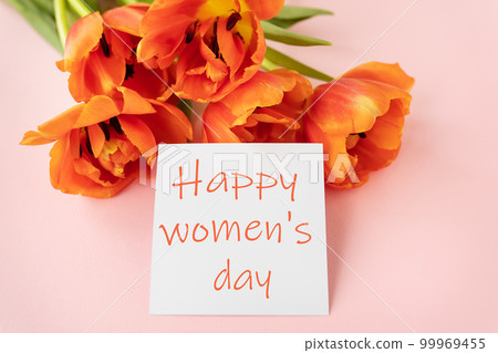 A beautiful bouquet of orange peony tulips along with a white card in the middle with the words happy women's day on a pink background. A beautiful bouquet of orange peony tulips along with a white card in the middle with the words happy women's day on a pink background. 99969455