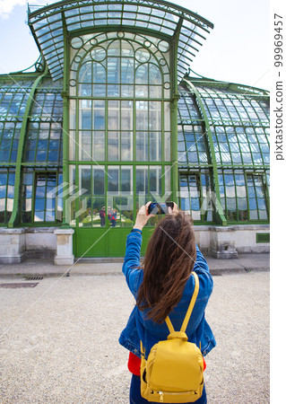 A young girl holds a phone in her hands and takes a picture of a beautiful building in Schonbrunn Park, Vienna. A young girl holds a phone in her hands and takes a picture of a beautiful building in Schonbrunn Park, Vienna. 99969457