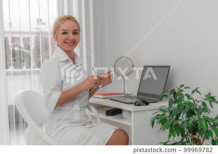 Blonde doctor in the doctor's office smiling. On the table is a computer, a mirror. The office has white walls, a flower with green leaves. Blonde doctor in the doctor's office smiling. On the table is a computer, a mirror. The office has white walls, a flower with green leaves. 99969782