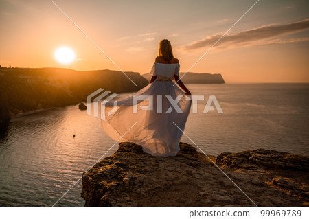 Sunset woman dress. A happy woman in a white light dress stands on the edge of a cliff above the sea, waving a white long dress, against the background of the sunrise of the sun and the sea. Sunset woman dress. A happy woman in a white light dress stands on the edge of a cliff above the sea, waving a white long dress, against the background of the sunrise of the sun and the sea. 99969789