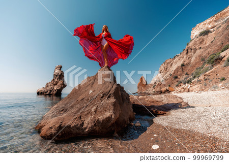 Beautiful woman posing in a luxurious red dress for the beach and in a white swimsuit with a long train, standing on a cliff against the sea. Around the cliff, the seashore. 99969799