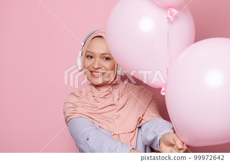 Beautiful Middle-Eastern Muslim woman wearing wireless headphones and pink hijab, holding out at camera a bunch of pastel helium party balloons, smiling looking at camera, isolated color background Beautiful Middle-Eastern Muslim woman wearing wireless headphones and pink hijab, holding out at camera a bunch of pastel helium party balloons, smiling looking at camera, isolated color background 99972642