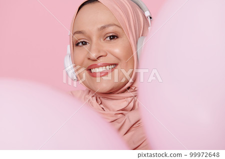 Close-up portrait of middle aged Muslim woman wearing headphones and pink hijab, smiling with beautiful toothy smile, peeking out from behind pink pastel inflatable balloons, isolated pink background Close-up portrait of middle aged Muslim woman wearing headphones and pink hijab, smiling with beautiful toothy smile, peeking out from behind pink pastel inflatable balloons, isolated pink background 99972648