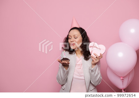 Attractive brunette woman making cherished wish and blowing out a candle on her birthday cake, holding a pink heart shaped gift box, posing with inflatable pink balloons, on isolated pink background 99972654