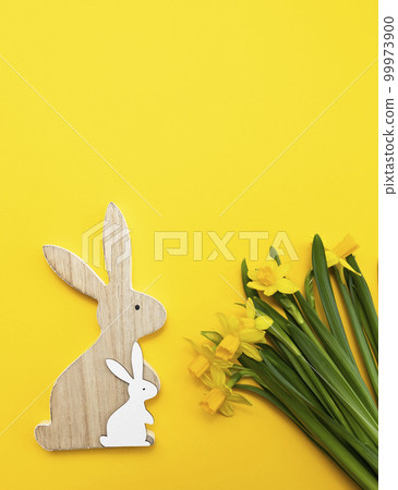 Wooden rabbit and bunny, yellow fresh narcissus flowers with green leaves on orange yellow background. Easter poster,celebration.Flat lay monochromatic greetings and presents for Easter Day,verticall 99973900