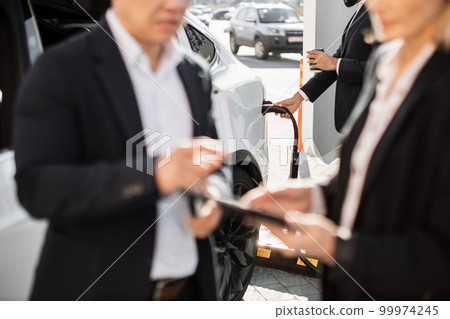 Business people using gadgets and charging electric car Business people using gadgets and charging electric car 99974245