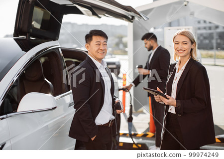 Multiracial colleagues standing at station and charging EV 99974249