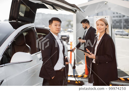 Multiracial colleagues standing at station and charging EV Multiracial colleagues standing at station and charging EV 99974250