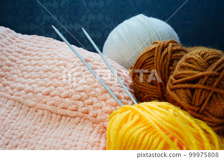 Two knitting needles stick out from a ball of yarn in the background of a piece of knitted goods. Yellow, coral pink, brown, white yarns of different thicknesses and compositions. Close-up, copy space Two knitting needles stick out from a ball of yarn in the background of a piece of knitted goods. Yellow, coral pink, brown, white yarns of different thicknesses and compositions. Close-up, copy space 99975808