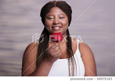 African american woman holding red rose love symbol portrait. Body positive smiling happy lady with flower showing beauty, passion and romance concept and looking at camera African american woman holding red rose love symbol portrait. Body positive smiling happy lady with flower showing beauty, passion and romance concept and looking at camera 99976165
