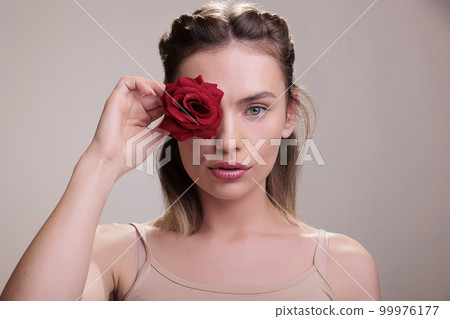 Beautiful young woman covering eye with red rose bud portrait. Attractive blonde fashion model wearing natural make up holding blooming flower near face and looking at camera Beautiful young woman covering eye with red rose bud portrait. Attractive blonde fashion model wearing natural make up holding blooming flower near face and looking at camera 99976177