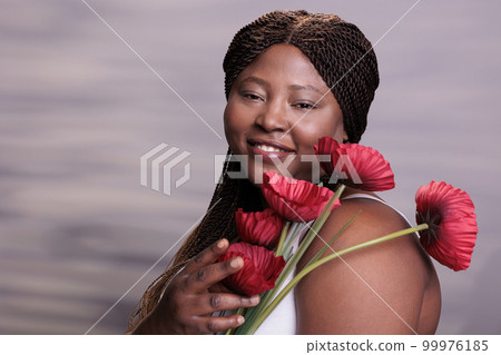 African american woman holding poppy flowers portrait, showing femininity and tenderness concept. Attractive plus size model posing with beautiful summer plants in studio close up 99976185