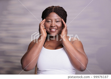 African american body positive model touching fresh healthy skin and looking at camera with happy facial expression. Beautiful plus size woman posing in studio for skincare cosmetology 99976187