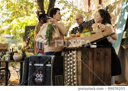 Happy delivery girl with thermo bag picking up order at farmers market. Female courier standing near farm produce stand taking fresh organic fruits and vegetables to deliver them to customer doorstep Happy delivery girl with thermo bag picking up order at farmers market. Female courier standing near farm produce stand taking fresh organic fruits and vegetables to deliver them to customer doorstep 99976203