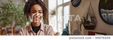Attractive smiling florist stands with crossed arms on background of her own floral decor studio Attractive smiling florist stands with crossed arms on background of her own floral decor studio 99976899
