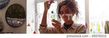 Smiling woman holding handmade glass decoration in the form of heart standing in home decor studio Smiling woman holding handmade glass decoration in the form of heart standing in home decor studio 99976910