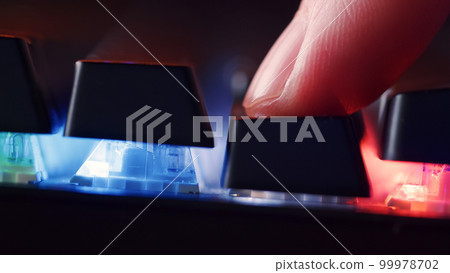 Human finger is clicking on the key on the computer keyboard, close up view. Finger presses  on the gaming keyboard key with led light, macro shot. Low angle view with a soft focus. 99978702