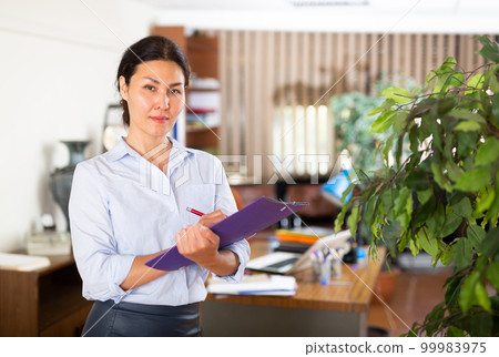 Friendly female administrative secretary standing in office with documents Friendly female administrative secretary standing in office with documents 99983975