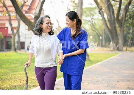 Lovely female caregiver assisting and supporting an old lady to walk with a walking stick 99988226