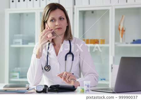 Female doctor calls her patient while sitting at table in medical clinic. Female doctor calls her patient while sitting at table in medical clinic. 99990037