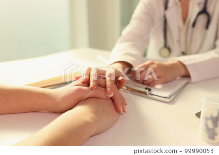 Doctor puts her hand on hands of patient, sitting with her in medical clinic. Doctor puts her hand on hands of patient, sitting with her in medical clinic. 99990053