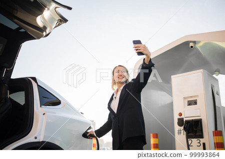 Woman having video call on mobile and charging electric car Woman having video call on mobile and charging electric car 99993864