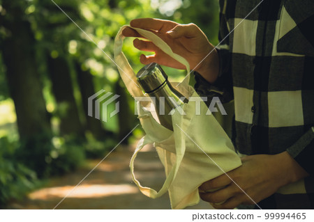 Close-up of Unrecognizable female hand holding Water bottle from eco bag. Reusable steel thermo water bottle in park. Sustainable lifestyle. Plastic free zero waste free living. Go green Woman 99994465