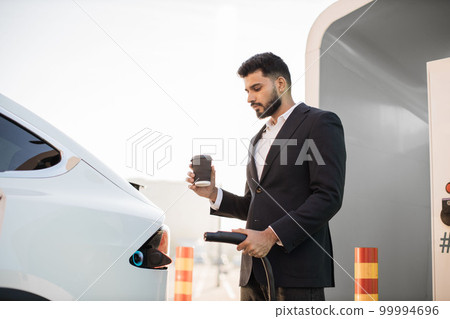 Businessman with coffee in hand charging modern car 99994696