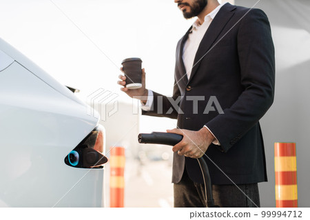 Close up of man holding coffee and charging electro car 99994712