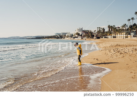 Boy in yellow rubber boots playing with water and sand at the beach. School kid touching water at autumn winter sea. Child having fun with waves at the shore. Spring Holiday vacation concept 99996250