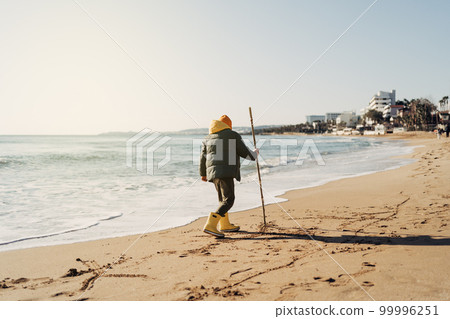 Boy in yellow rubber boots playing with stick and sand at the beach. School kid touching water at autumn winter sea. Child having fun with waves at the shore. Spring Holiday vacation concept 99996251