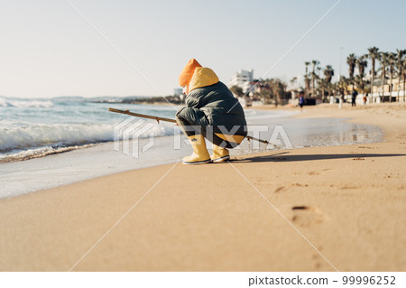 Boy in yellow rubber boots playing with stick and sand at the beach. School kid touching water at autumn winter sea. Child having fun with waves at the shore. Spring Holiday vacation concept 99996252
