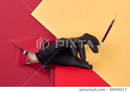 Hand with black glove holding cosmetic syringe with purple glitter. Red and yellow paper background. 99999017