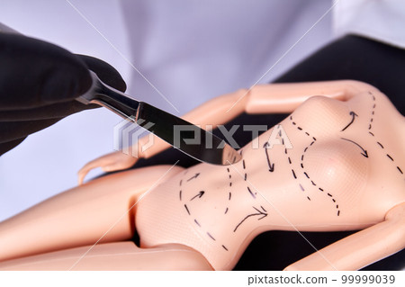 Close up surgeon cutting plastic dolls body. Metallic scalpel cutting belly along dotted line. 99999039