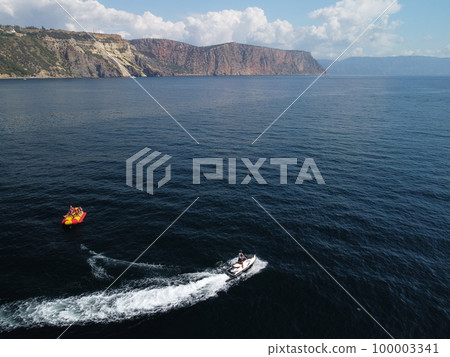 Happy people swim on air mattress behind a high speed water bike. Tourists ride on inflatable watercraft boat. Sea attraction. Water scooter skating people on large Inflatable raft. Aerial copter view 100003341