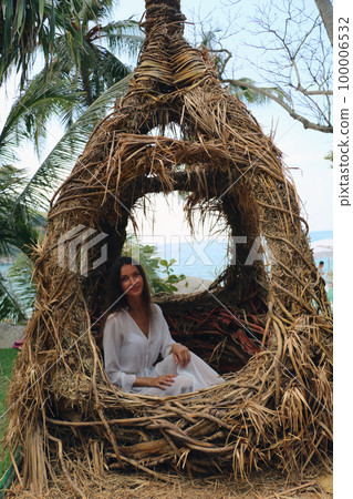 Beautiful young woman in a white beach cape sits in a large nest against a background of palm trees and the sea. 100006532