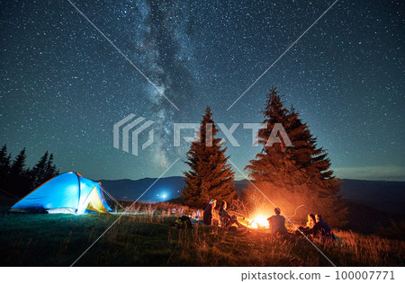 Night camping in mountains under starry sky. Group of people tourists having a rest near campsite, burning campfire and illuminated tent. Concept of tourism, hiking and adventure. 100007771