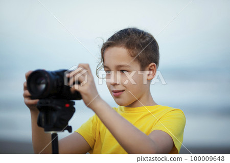 Boy teenager with digital camera on tripod outdoors. Child interested in photography-videography learning to take picture and make videos Boy teenager with digital camera on tripod outdoors. Child interested in photography-videography learning to take picture and make videos 100009648