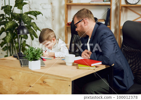 Mature man, father sitting at table, working remotely at home and doing homework with his little son 100012378
