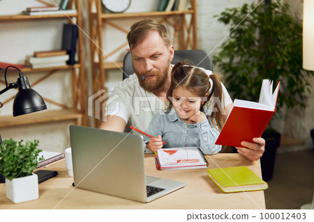 Mature man, father sitting at table and working on laptop remotely at home with his little preschool daughter sitting with him Mature man, father sitting at table and working on laptop remotely at home with his little preschool daughter sitting with him 100012403
