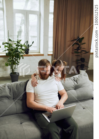 Mature man, father sitting on sofa and working on laptop remotely at home with his little daughter playing with him Mature man, father sitting on sofa and working on laptop remotely at home with his little daughter playing with him 100012525