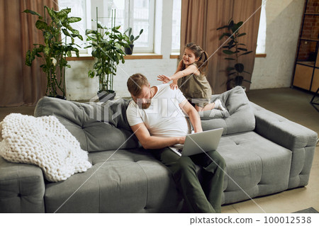 Mature man sitting on sofa in living room at home and working remotely. Little girl, daughter playing and distracting him Mature man sitting on sofa in living room at home and working remotely. Little girl, daughter playing and distracting him 100012538