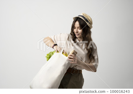 Smiling young woman in beige t-shirt and a hst with a mesh eco bag full of vegetables and on a white studio background. Sustainable lifestyle. Eco friendly concept. Zero waste. Smiling young woman in beige t-shirt and a hst with a mesh eco bag full of vegetables and on a white studio background. Sustainable lifestyle. Eco friendly concept. Zero waste. 100016243
