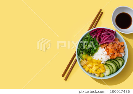 Poke bowl with rice, salmon,cucumber,mango,onion,wakame salad, poppy seeds ands sunflowers seeds on yellow background 100017486