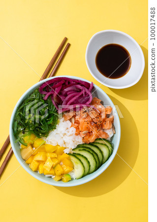 Poke bowl with rice, salmon,cucumber,mango,onion,wakame salad, poppy seeds ands sunflowers seeds on yellow background 100017488