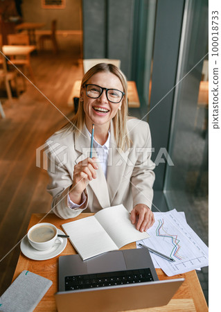 Smiling businesswoman drinking coffee and making notes while working on laptop in cafe Smiling businesswoman drinking coffee and making notes while working on laptop in cafe 100018733