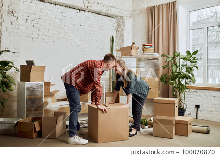 Happy young family, man and woman moving into new house, apartment. Opening many cardboard boxes. Furnishing room Happy young family, man and woman moving into new house, apartment. Opening many cardboard boxes. Furnishing room 100020870